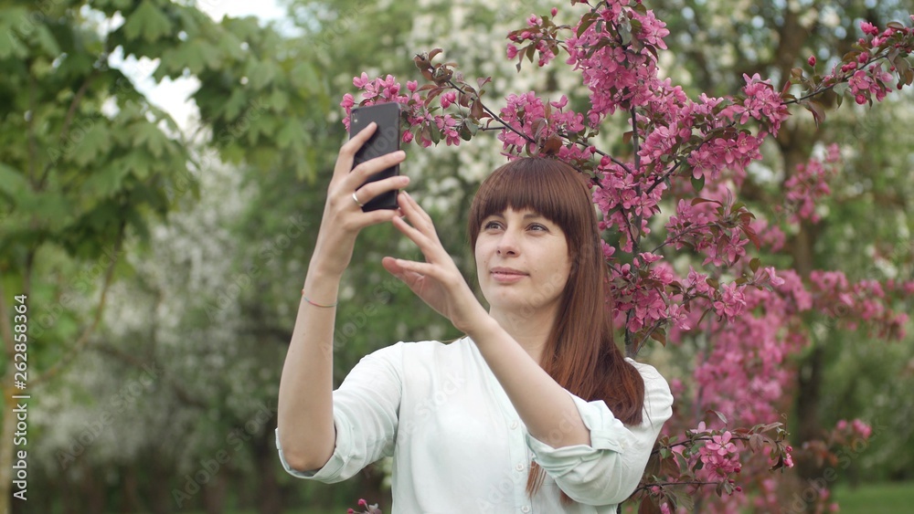 Girl makes selfie in the garden. An attractive red-haired woman smiles making selfi using a mobile phone in a cherry orchard. The concept of using gadgets for a healthy lifestyle. Girl makes selfie in the garden. An attractive red-haired woman smiles making selfi using a mobile phone in a cherry orchard. The concept of using gadgets for a healthy lifestyle.