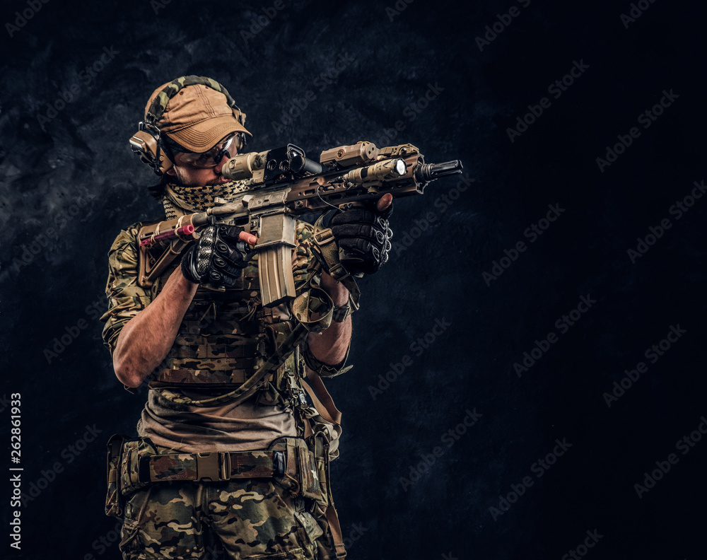 The elite unit, special forces soldier in camouflage uniform holding an assault rifle with a laser sight and aims at the target. Studio photo against a dark wall The elite unit, special forces soldier in camouflage uniform holding an assault rifle with a laser sight and aims at the target. Studio photo against a dark wall