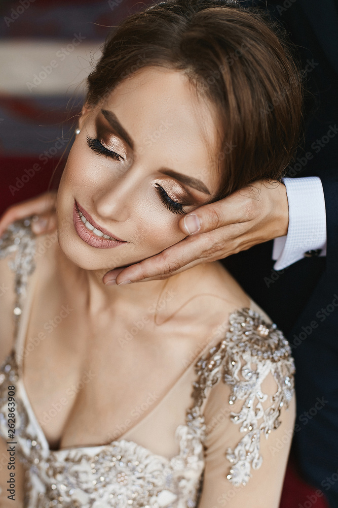 Stylish and beautiful brown-haired model girl with wedding hairstyle and bright makeup in stylish lace dress lean on male hand