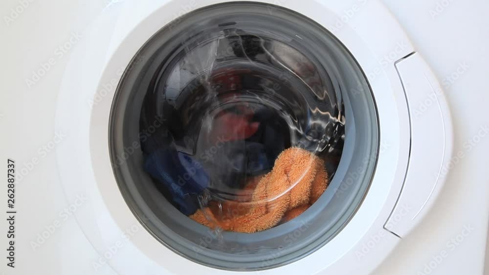 Sound included. Six second video of the start of the wash, the hiss of water entering the machine bowl running across the glass. Includes orange towel, white front load washing machine.