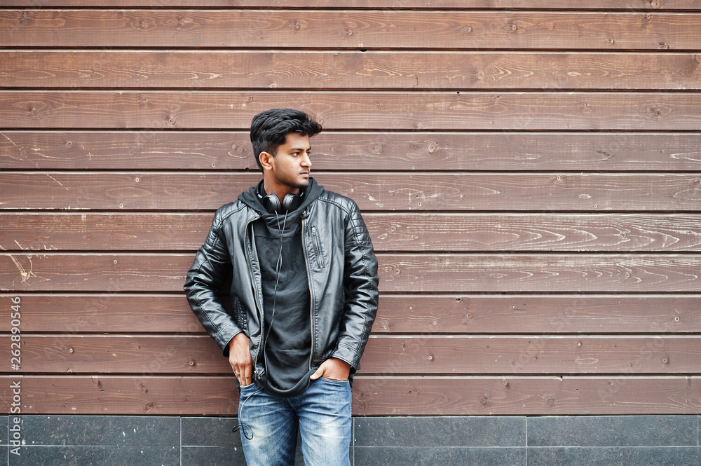 Stylish and casual asian man in black leather jacket, headphones posed against wooden wall.