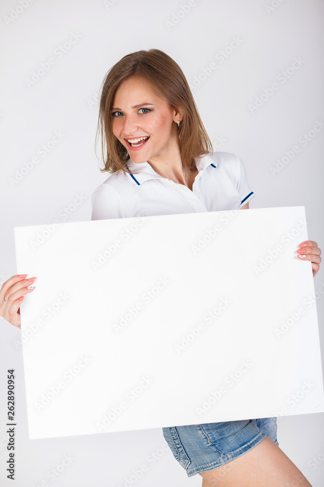 Beautiful young gir in denim shorts and a T-shirth on a white background holding big white blank card. Pretty student girl having fun and holding white empty signboard background with copy space. Beautiful young gir in denim shorts and a T-shirth on a white background holding big white blank card. Pretty student girl having fun and holding white empty signboard background with copy space.