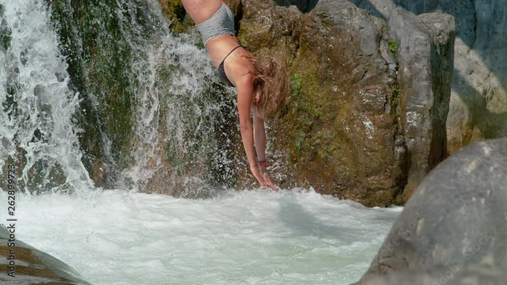 SLOW MOTION: Athletic tourist girl dives head first into a pond under the refreshing glassy waterfall. Sporty Caucasian woman diving off the edge of a waterfall and into the foaming stream water.