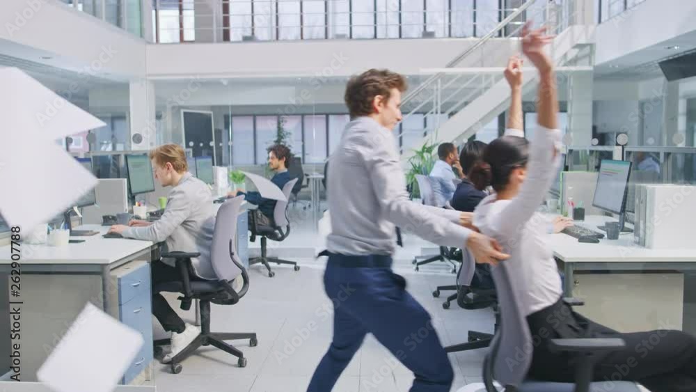 Cheerful Employee Pushes His Beautiful Colleague on a Chair Next to Diverse Business People Working on Computers in Modern Office Space. Female Throws Business Papers and Contracts in the Air. Cheerful Employee Pushes His Beautiful Colleague on a Chair Next to Diverse Business People Working on Computers in Modern Office Space. Female Throws Business Papers and Contracts in the Air.