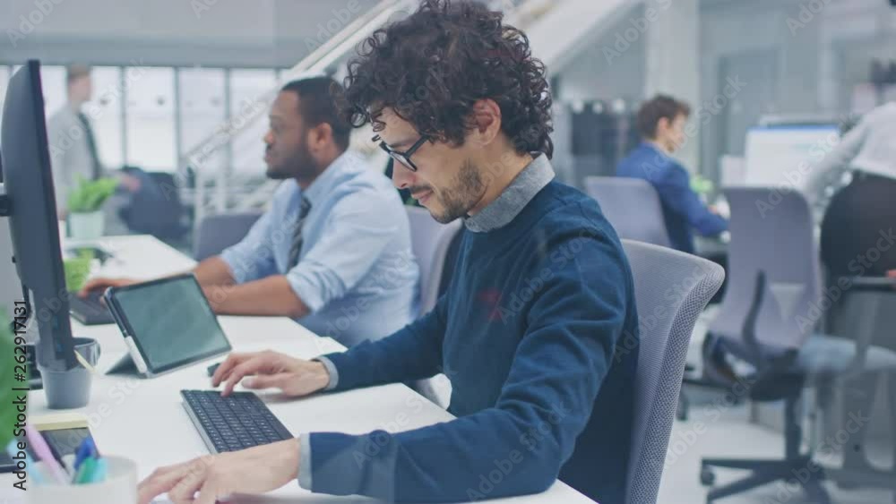 Young Handsome Manager with Curly Hair Works on a Desktop Computer and Compares Data with a Software. Diverse and Motivated Business People Work on Computers in Modern Open Office.