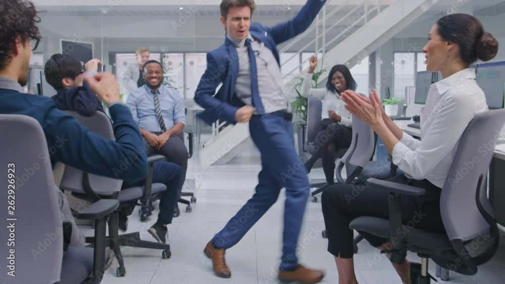Young Happy Business Manager Wearing a Suit and Tie Dancing and Giving High Fives in the Office. Celebrating Success. Diverse and Motivated Business People Work on Computers in Modern Open Office. Young Happy Business Manager Wearing a Suit and Tie Dancing and Giving High Fives in the Office. Celebrating Success. Diverse and Motivated Business People Work on Computers in Modern Open Office.
