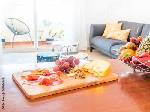 Table with fruit and food in a stylish bright living room with sofa, wooden table, side tables, tv,plants and big window to a terrace with mountain view. Healthy lifestyle concept.