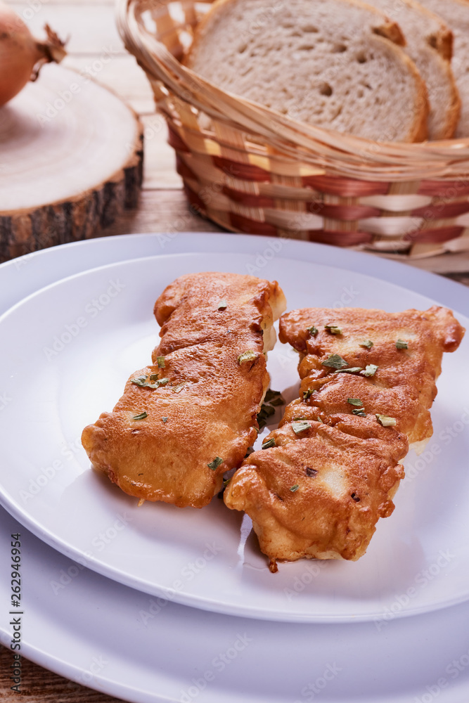 Deep-fried battered cod fillets, in a white plate next to onions and bread on a wooden background. Close-up