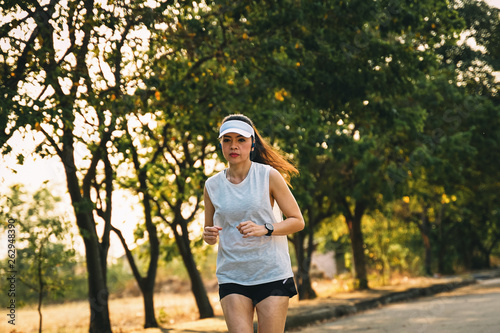 Young woman runner wears white sport vest, white cap, black short pant runs on the road to exercise in the evening while listens to music through earphone. Female runner runs on the city road.