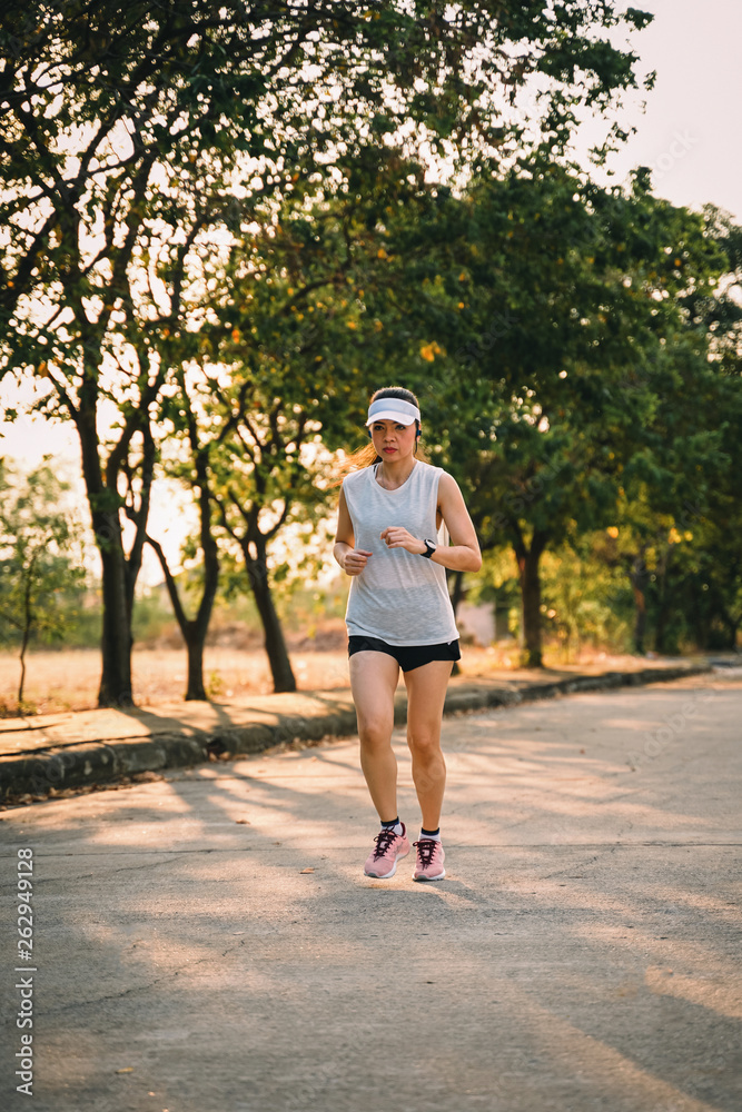 Fototapeta premium Young woman runner wears white sport vest, white cap, black short pant runs on the road to exercise in the evening while listens to music through earphone. Female runner runs on the city road.
