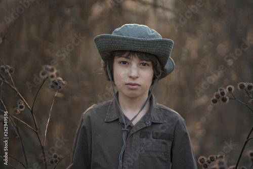 A boy in a hat and coveralls is standing in dry bushes of burdock.