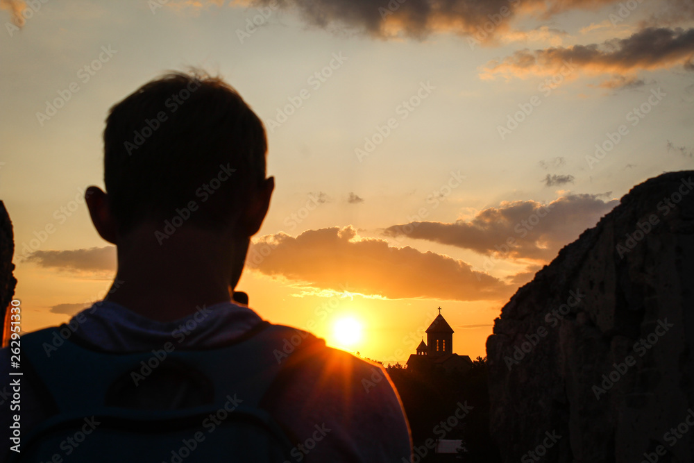 A man with a blue backpack on his shoulders makes a photo of a sunset on a mobile phone. City, church in the rays of the setting sun. Golden light. Beautiful sky with clouds. Georgia. Tourism, travel