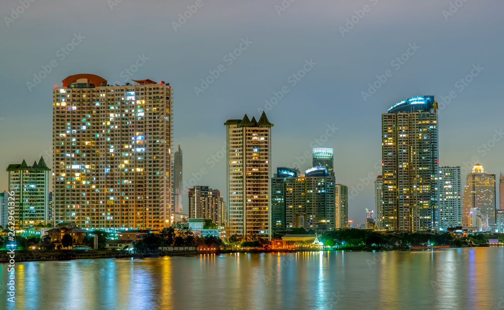 Naklejka premium Cityscape of modern building near the river in the night. Modern architecture office building. Skyscraper with evening sky. Night photography of riverfront building. Condominium open light in night.
