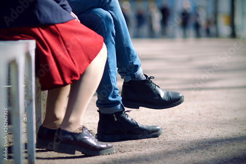 Legs of a young couple sitting on a summer day on a bench. The man is dressed in tight jeans and black boots, and the woman in a red skirt and Burgundy shoes.