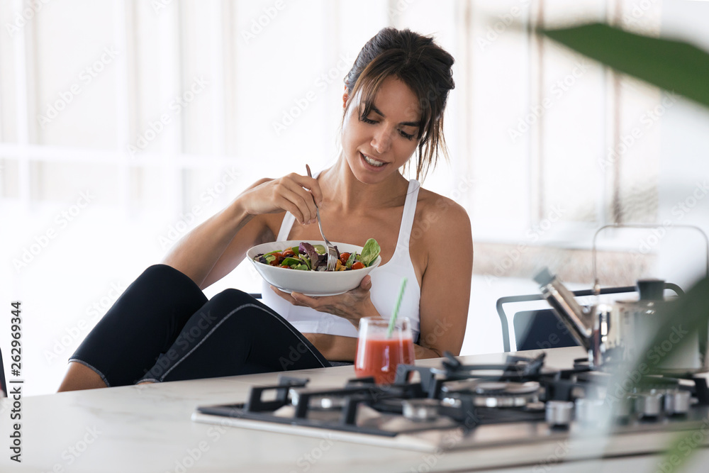 Sporty young woman eating salad and drinking fruit juice in the kitchen at home. - obrazy, fototapety, plakaty 