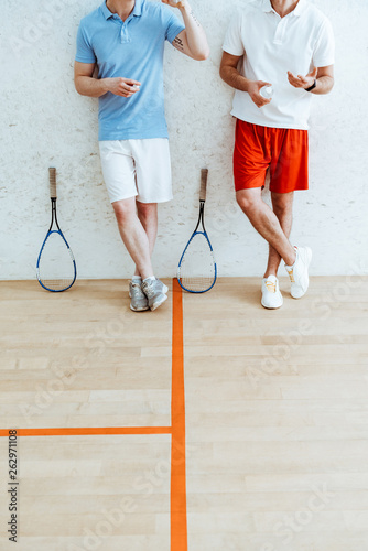 Cropped view of two squash players in shorts standing with crossed legs in four-walled court