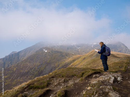 A lone walker looks at a map on the way to the misty summit of Blencathra (also known as Saddleback) in the Lake District, Cumbria, UK with cloud falling against a blue sky