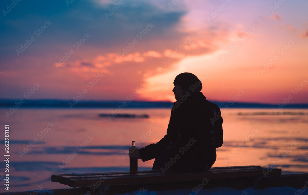 An abandoned lonely guy sits on a bench and wants to drink a bottle of beer. Male alcoholism. Depression and loneliness. Alcohol and concept. Beautiful photo at sunset. Evening winter silhouette.