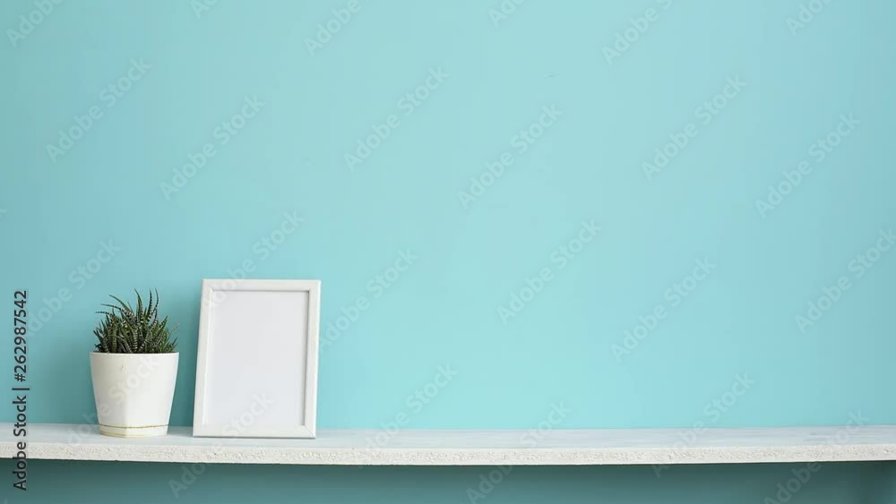 Modern room decoration with Picture frame mockup. White shelf against pastel turquoise wall with hand putting down potted succulent plant.