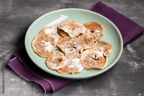 Apple chips with powdered sugar and cinnamon on green plate on purple fabric on grey background. 