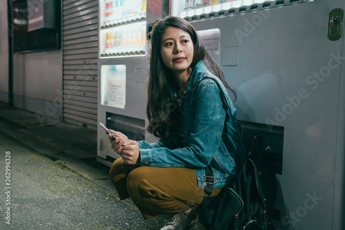 young relaxing female backpacker kneeling down rely on vending machine by dark street side at night in osaka japan. beautiful woman waiting for someone resting by auto vendor holding cellphone photo