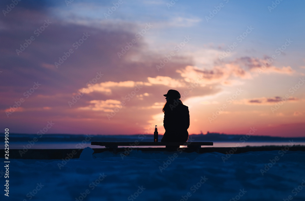 A young lonely girl sits on a bench and is sad next to a bottle of beer. Female alcoholism. Depression and loneliness. Alcohol and concept. Beautiful photo at sunset. Evening winter silhouette. A young lonely girl sits on a bench and is sad next to a bottle of beer. Female alcoholism. Depression and loneliness. Alcohol and concept. Beautiful photo at sunset. Evening winter silhouette.