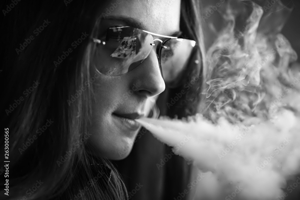 Fototapeta premium Vape teenager with problem skin. Portrait of young cute girl in sunglasses smoking an electronic cigarette in the bar. Bad habit that is harmful to health. Vaping activity. Black nad white.