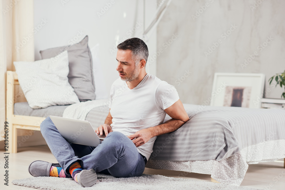 Handsome middle aged man sitting on the floor wirh crossed legs, leaning on bed and using laptop.