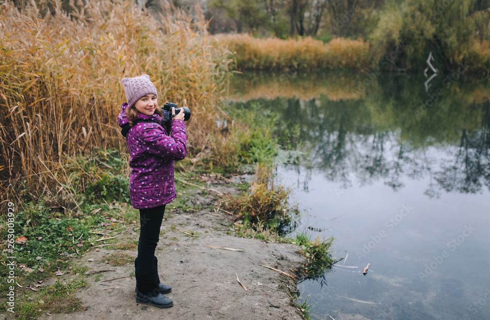 A young girl in a purple jacket and hat walks with a camera on the nature in the fall near the lake. Baby photographer. Happy childhood. Portrait and autumn photography. A young girl in a purple jacket and hat walks with a camera on the nature in the fall near the lake. Baby photographer. Happy childhood. Portrait and autumn photography.