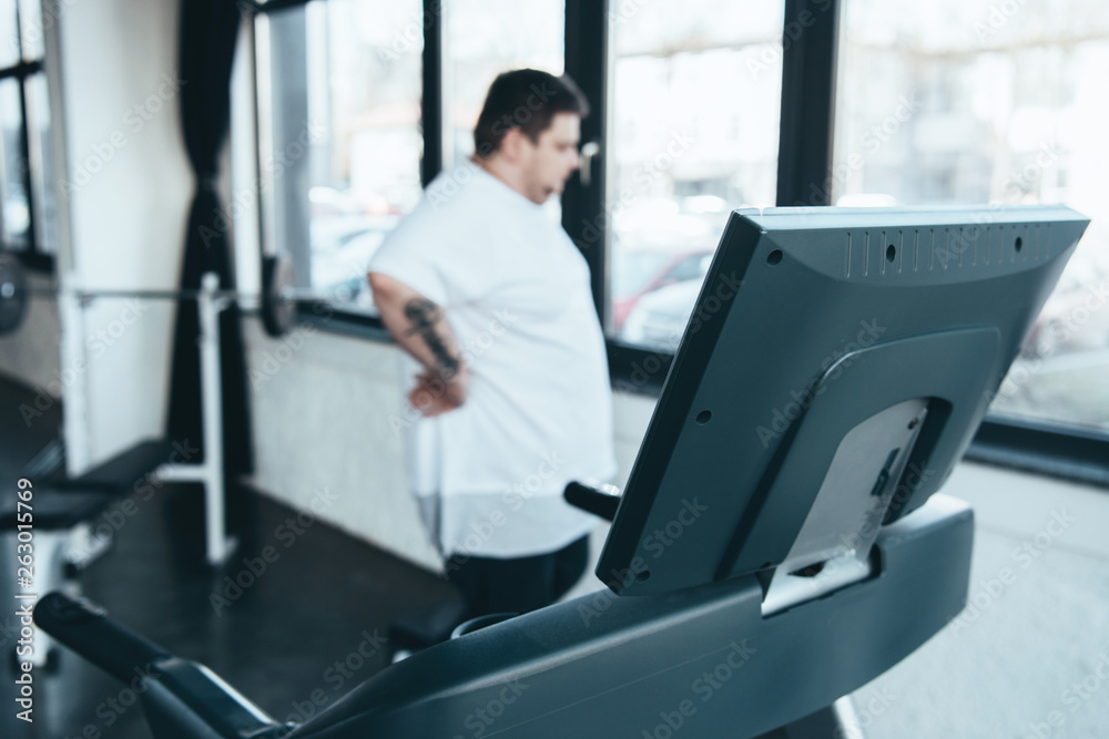selective focus of treadmill and overweight man looking through window at sports center selective focus of treadmill and overweight man looking through window at sports center