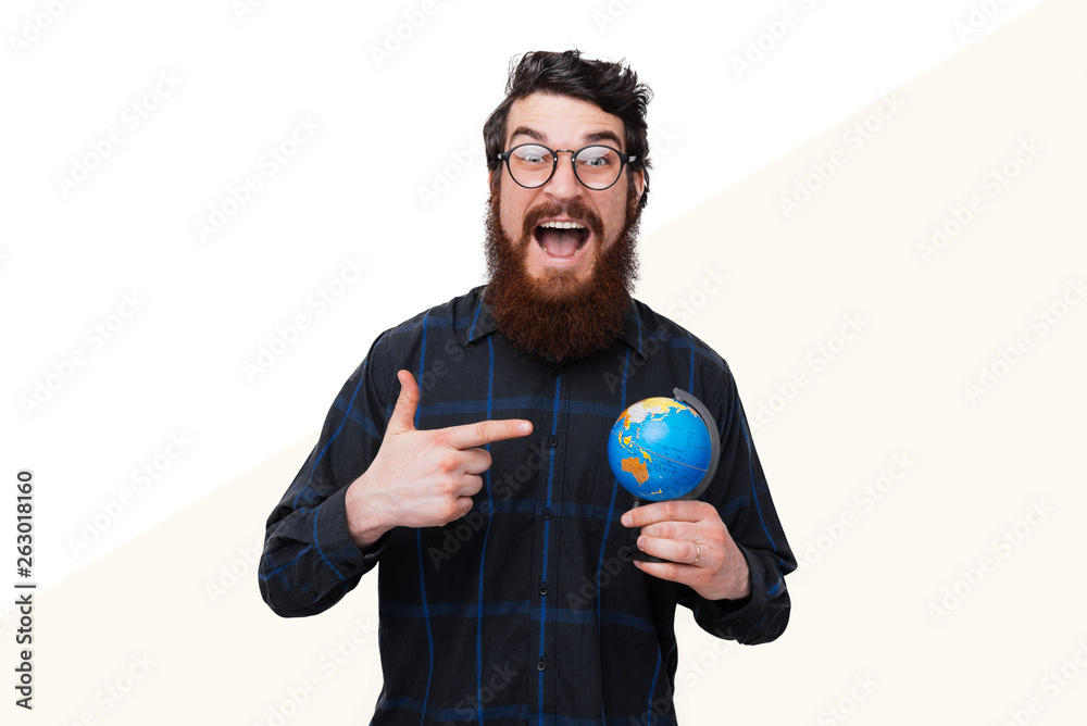 Young excited bearded man wearing glasses and holding small Earth globe and pointing to it. Young excited bearded man wearing glasses and holding small Earth globe and pointing to it.