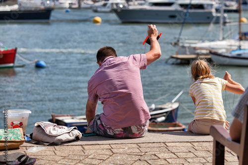 A father and daughter crabbing over the side of a harbour in the summer with sailboats in the background
