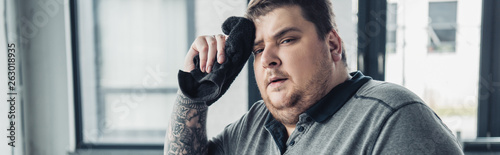 panoramic shot of tired overweight man looking at camera and wiping face with towel at sports center