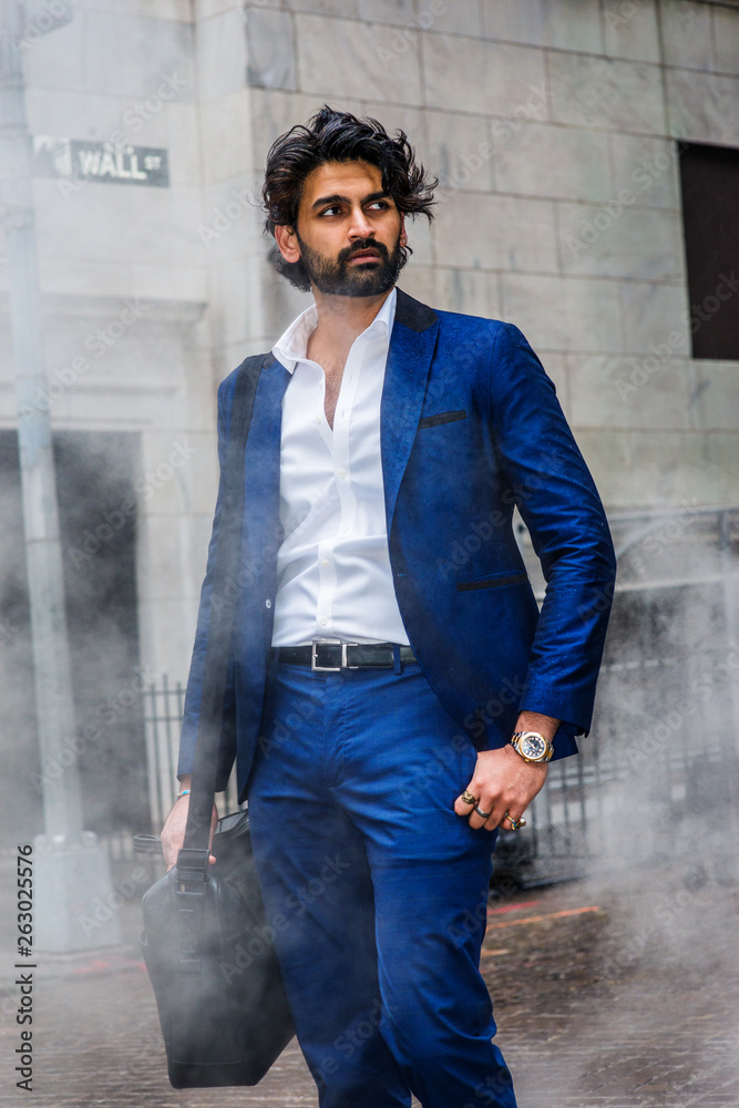 Obraz premium Raining day - grainy, foggy, wet feel. Young East Indian American Man with beard traveling in N, wearing blue suit, white shirt, carrying shoulder bag, looking away, walking on Wall Street in New York