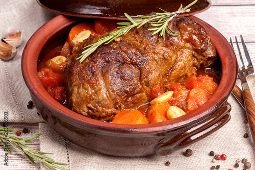 A large piece of meat baked with vegetables and herbs on an earthenware plate. Food for ketogenic diet. Cooked in the home kitchen. Wooden table and natural napkin. Horizontal view.