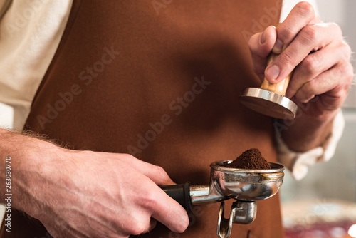 Cropped view of barista in brown apron holding portafilter with ground coffee and tamper