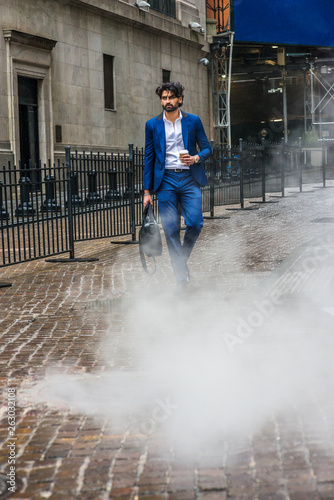 Raining day - grainy, foggy, wet feel. Young East Indian American Business Man with beard, wearing blue suit, white shirt, carrying leather hand bag, holding coffee cup, walking on street in New York