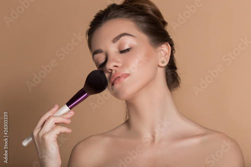 Closeup portrait of beautiful girl with perfect skin is holding makeup brush in hand. Isolated on beige. Beauty and cosmetics concept.