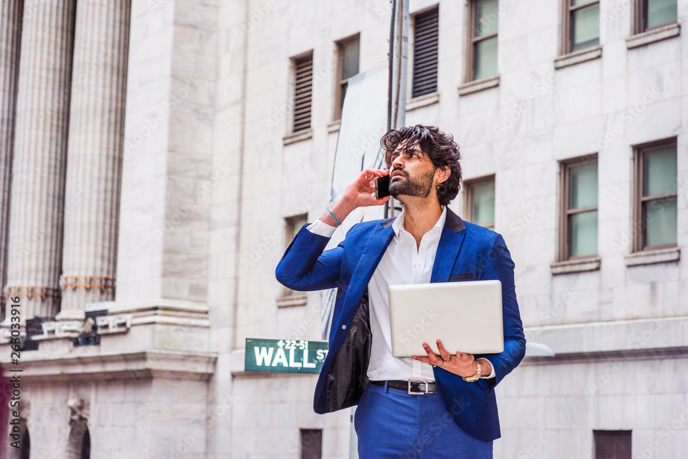 Way to Success. Young East Indian American Businessman with beard traveling, working in New York, wearing blue suit, holding laptop computer, standing on Wall Street, looking up, talking on cell phone