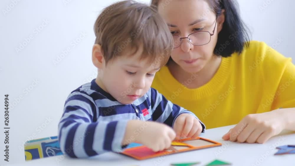 little asian boy collects puzzles with mom sitting at the desk. A cute child collects a picture from large multi-colored details. Playing with a puzzle. slow motion. Teacher playing with boy little asian boy collects puzzles with mom sitting at the desk. A cute child collects a picture from large multi-colored details. Playing with a puzzle. slow motion. Teacher playing with boy