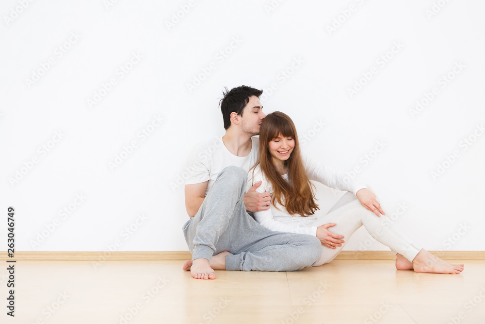 Happy family at home. Beautiful young couple in white pajamas kisses and hugs sitting on the floor during weekends at home. Love concept, white background Happy family at home. Beautiful young couple in white pajamas kisses and hugs sitting on the floor during weekends at home. Love concept, white background