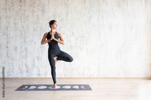 Obraz Beautiful young brunette woman yoga instructor doing vrikshasana on a mat in a wooden floor standing in the gym with day lighting
