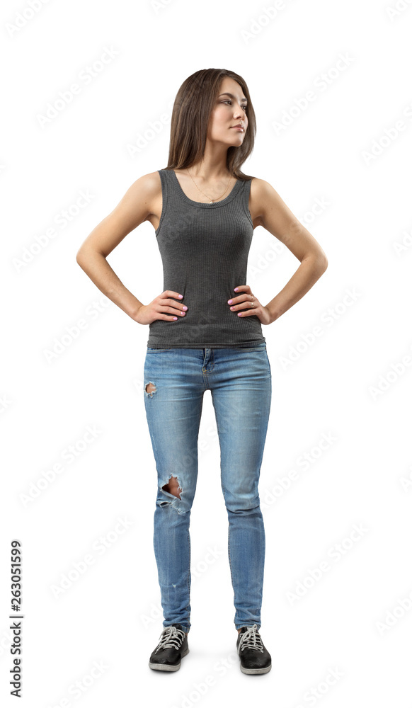 Front view of young attractive woman in gray sleeveless top and blue jeans standing and looking away with hands on hips isolated on white background.