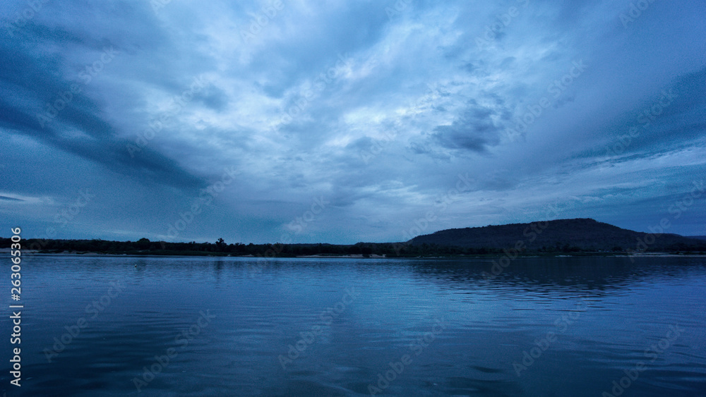 Blue sky with dark clouds with silhouette of mountain and tree at horizontal line with light reflection in the river