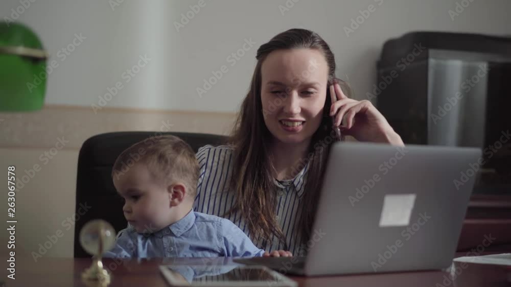 Portrait of busy pretty young mother working at home with the baby on her laps. The woman talking by cellphone while her son playing with small ball and touching keyboard on laptop. Freelance concept Portrait of busy pretty young mother working at home with the baby on her laps. The woman talking by cellphone while her son playing with small ball and touching keyboard on laptop. Freelance concept
