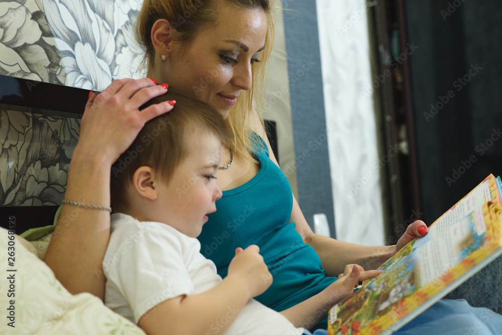 Obraz premium Young happy mom and little son are reading a book in bed