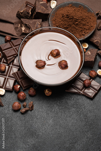 glass bowl of chocolate cream or melted chocolate, pieces of chocolate and hazelnuts on dark concrete background