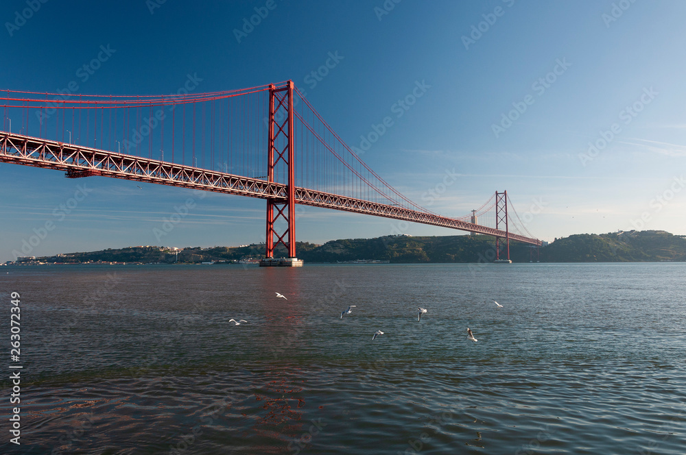 View of the 25 of April Bridge (Ponde 25 de Abril) with seagulls flying over the Tagus River, in the city of Lisbon, Portugal View of the 25 of April Bridge (Ponde 25 de Abril) with seagulls flying over the Tagus River, in the city of Lisbon, Portugal