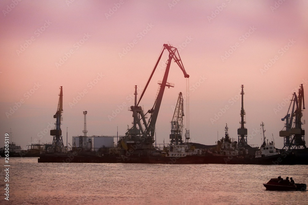 Port docks at sunset. Unloading ships on background of evening sky. Sea port and unloading of cargo containers. Silhouette of dock cranes in port and boat on the water