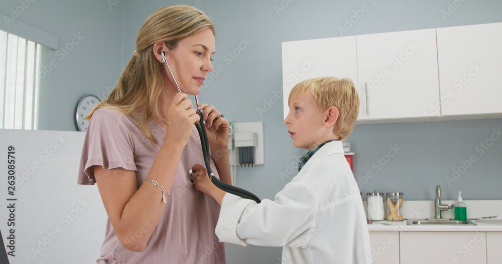 Cute child pretending to be a doctor with his mother while in pediatricians office using stethoscope. Little boy wearing oversized lab coat in exam room Cute child pretending to be a doctor with his mother while in pediatricians office using stethoscope. Little boy wearing oversized lab coat in exam room